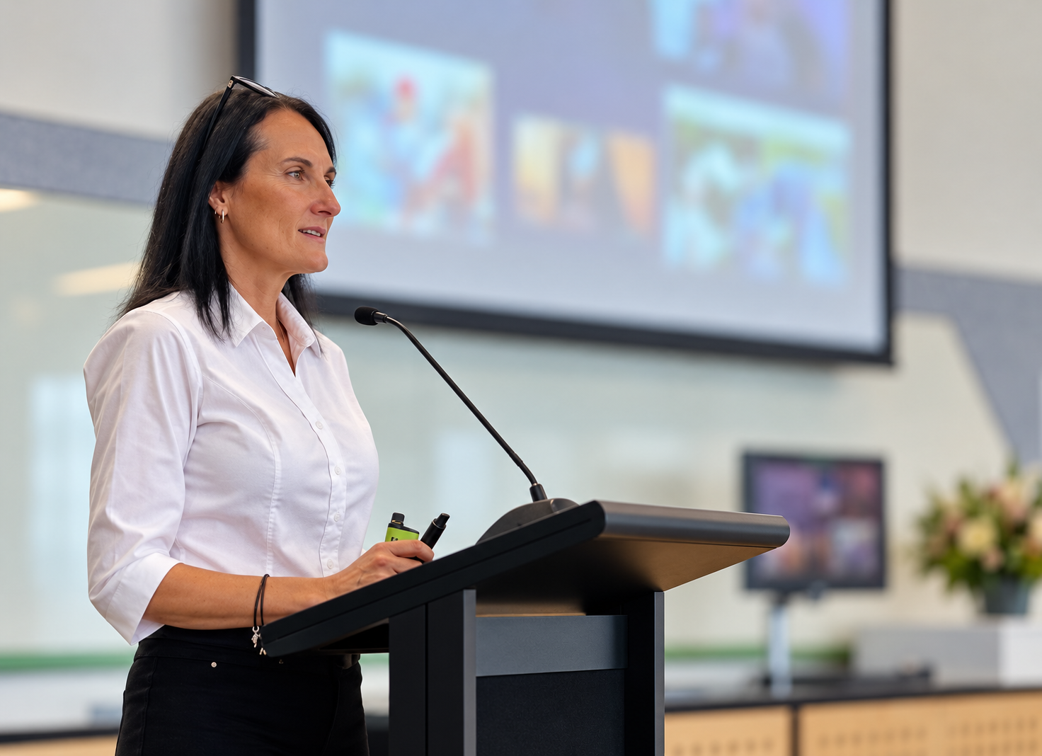 A woman with dark hair and a white shirt stands at a podium, giving a presentation in a conference room. She holds a clicker in her right hand, and there is a microphone attached to the podium. Behind her is a large screen displaying a slide with var