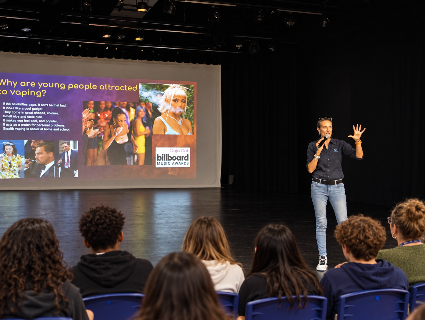 A woman giving a presentation on stage with a microphone, in front of a large screen displaying a slide titled 'Why are young people attracted to vaping?' The slide includes images of young people and celebrities, with a mix of text and logos. The au