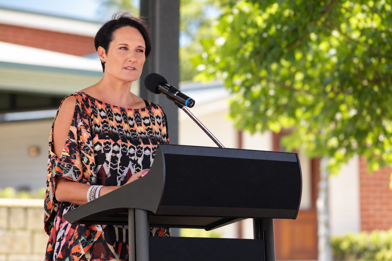 A woman with short dark hair speaking at a podium outdoors with a microphone, wearing a patterned dress with cut-out shoulders, and standing in front of green trees and buildings.