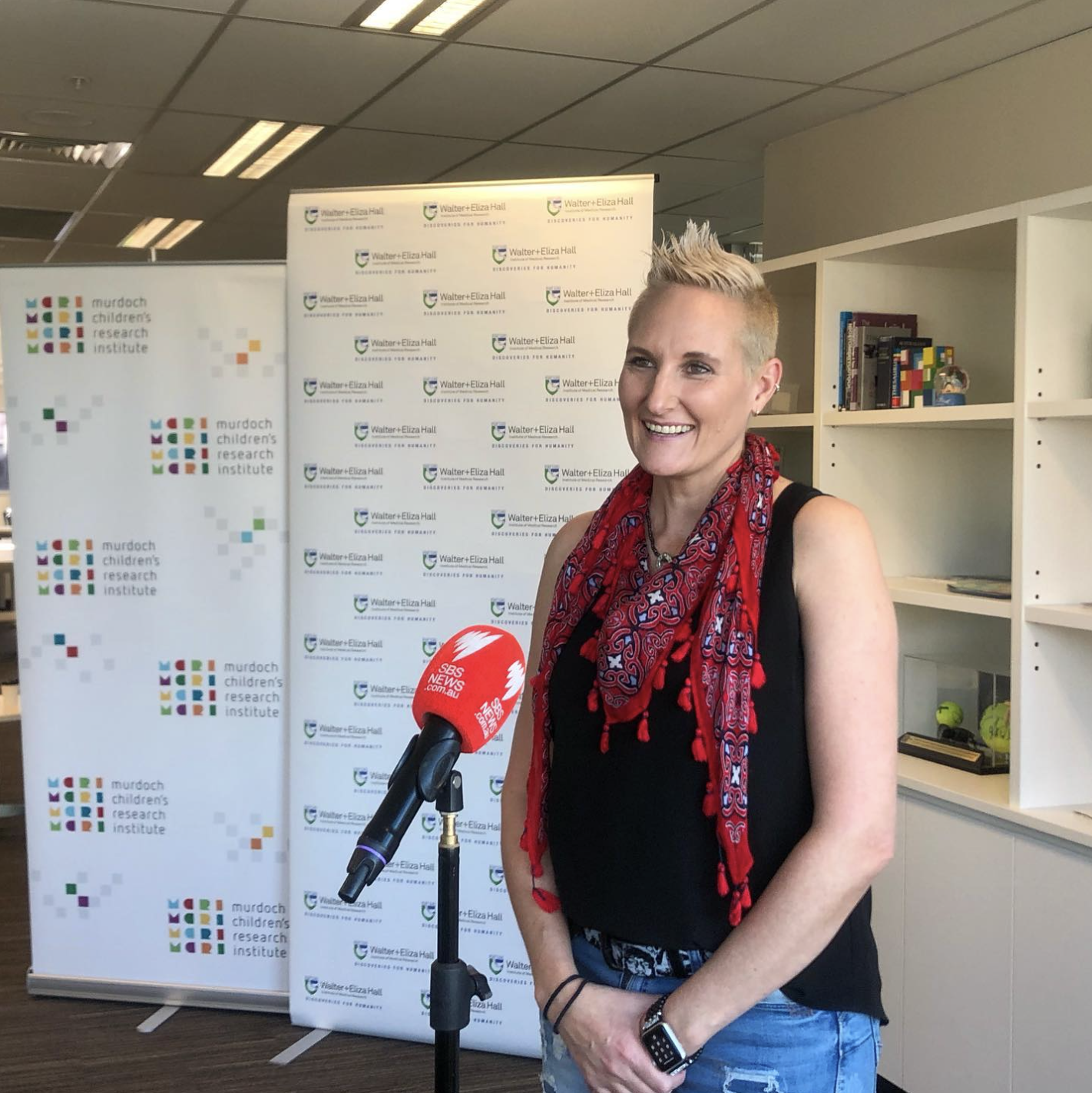 Woman being interviewed with a red SBS News microphone, standing in front of banners for Murdoch Children's Research Institute and Walter+Eliza Hall.