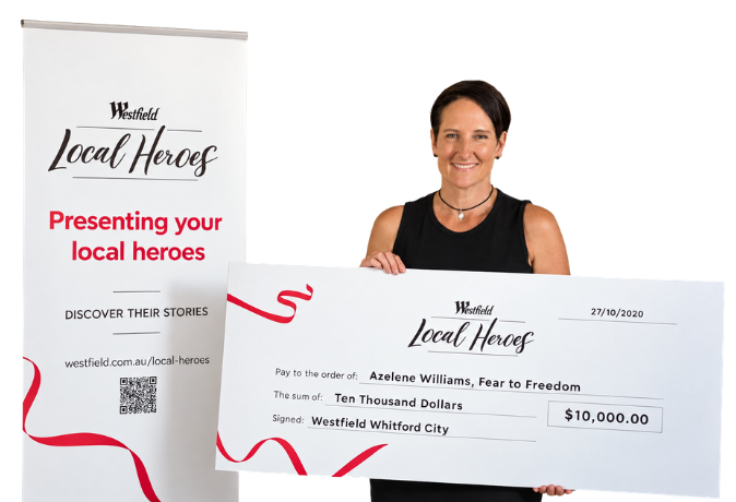 Azelene Williams holding a large check for ten thousand dollars as part of a local heroes recognition event, standing next to a promotional banner.