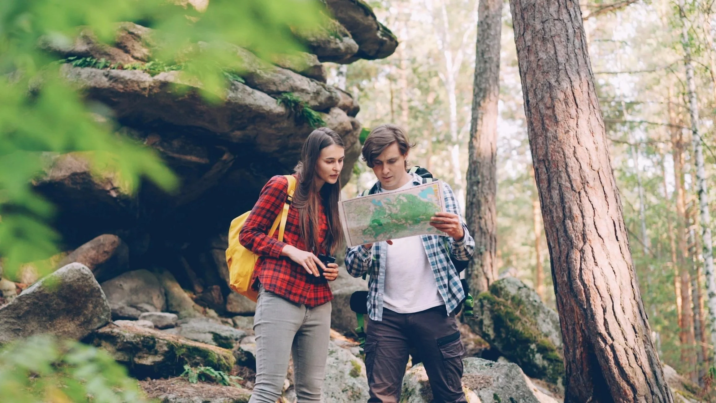 A young man and woman in a forest looking at a map, both carrying backpacks.