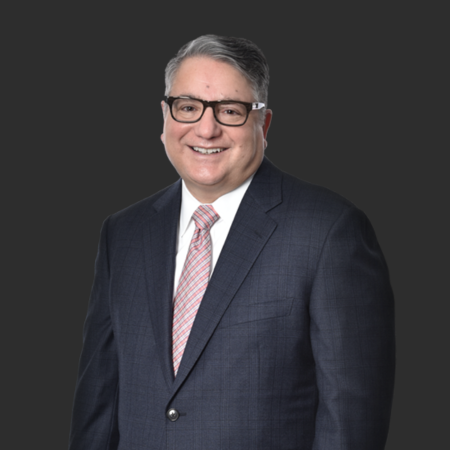 A smiling man with glasses in a navy suit with a patterned tie, standing against a dark gray background.