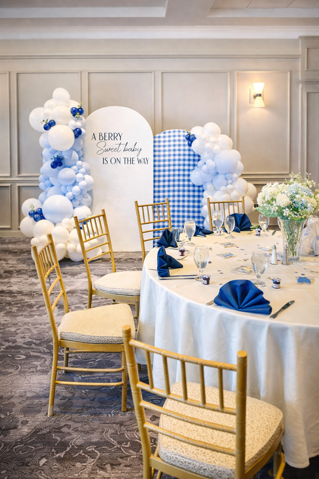 Baby shower event setup featuring a custom balloon installation, blue gingham backdrop, and styled round tables with gold chairs and navy napkins.