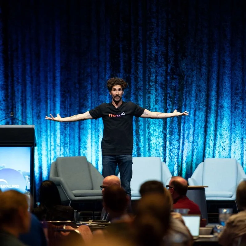 Gus Balbontin on stage with arms outstretched mid-keynote, lit by a blue backdrop, addressing an audience seated at tables in the foreground.