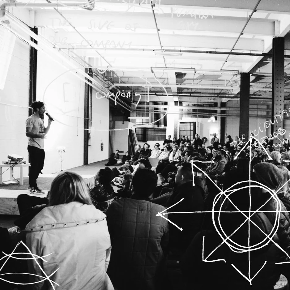 Gus Balbontin on stage with a microphone addressing a seated audience in an industrial venue, black and white photo with hand-drawn arrow and compass graphics.