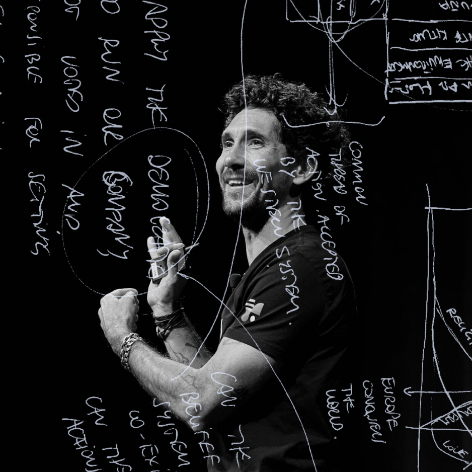 Gus Balbontin smiling mid-keynote and holding a presentation clicker, with handwritten notes and diagrams overlaid across a black background, in black and white.