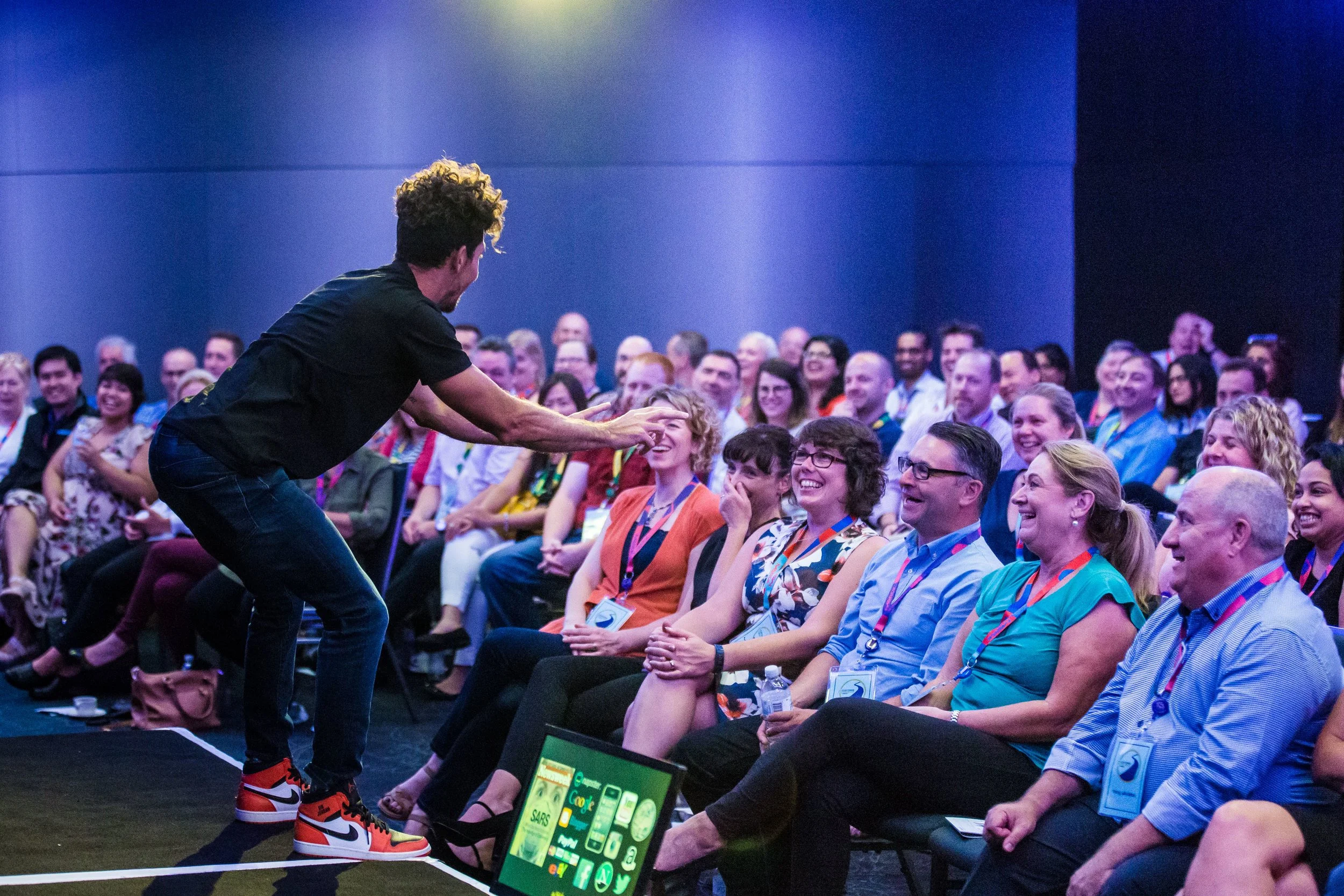 Gus Balbontin, a man with curly hair and a black shirt is engaging with an audience at a conference, with several people smiling, laughing, and wearing conference badges, sitting in dark chairs on a dark-colored carpeted floor.