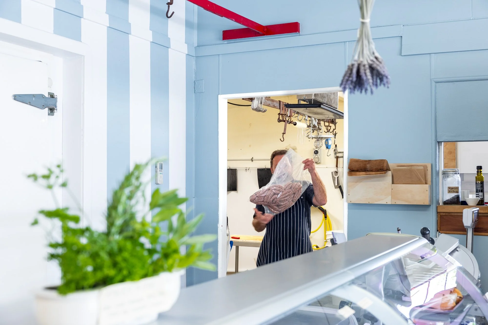 T Dunn, the Akaroa Butcher in a striped apron holding a large plastic bag of meat in a butcher shop or meat market, with tools and shelves in the background.