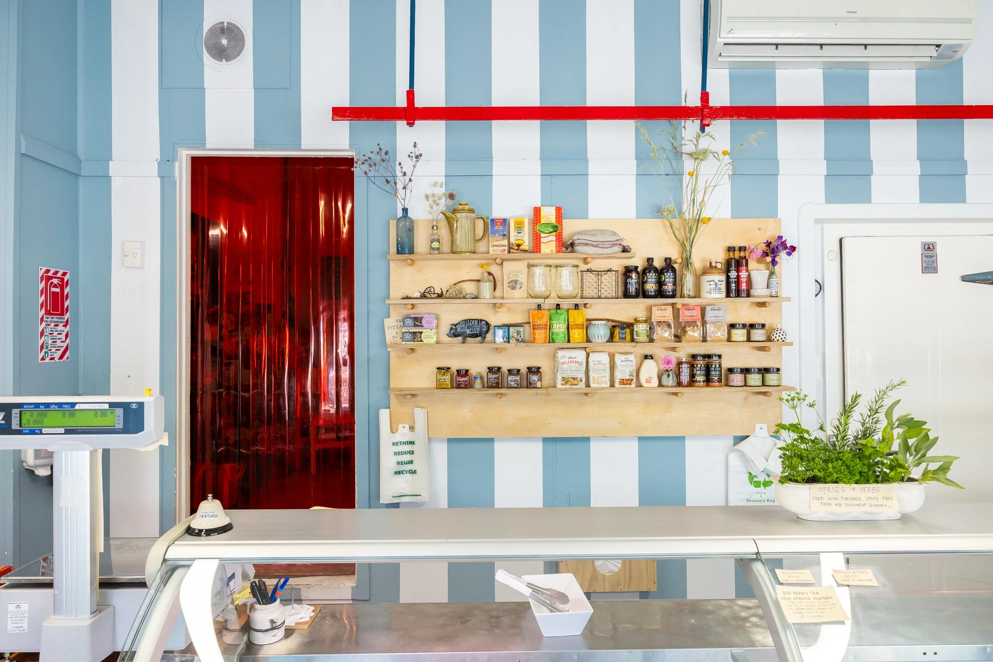 Interior of the Akaroa Butchery with light blue vertical striped walls, a wooden wall shelf holding various jars and bottles, a plant in a white pot, a red door with a reflective surface, and a white countertop with a cash register and scale.