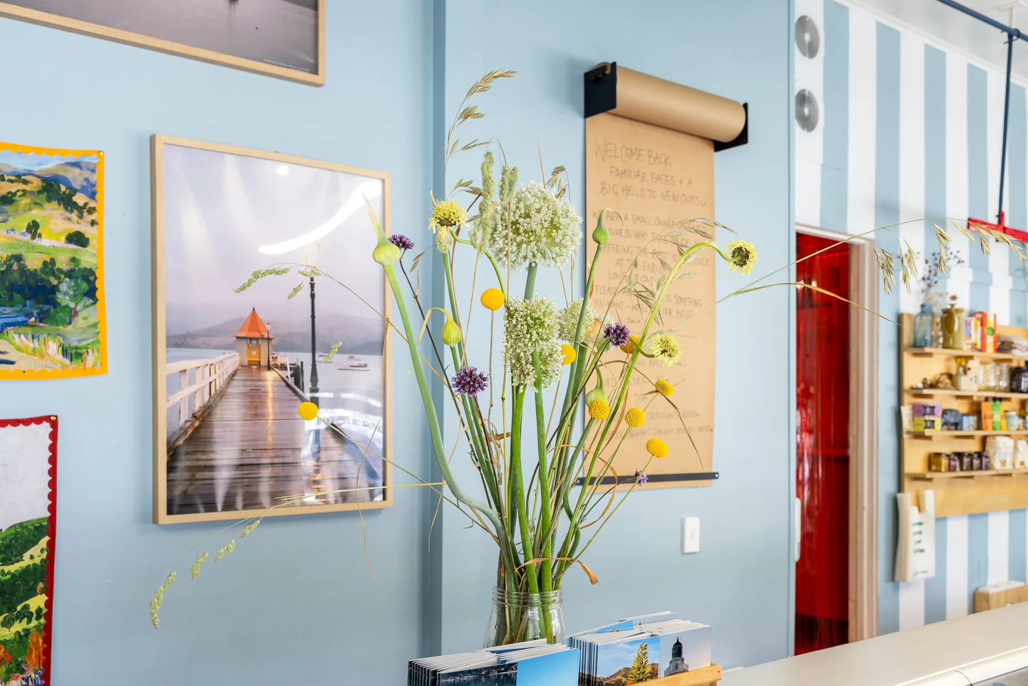 Inside the Akaroa and Butchery - Vase with a variety of wildflowers including yellow billy balls, green and white Queen Anne's lace, purple allium, and tall grass, placed on a counter in a bright, colorful room.