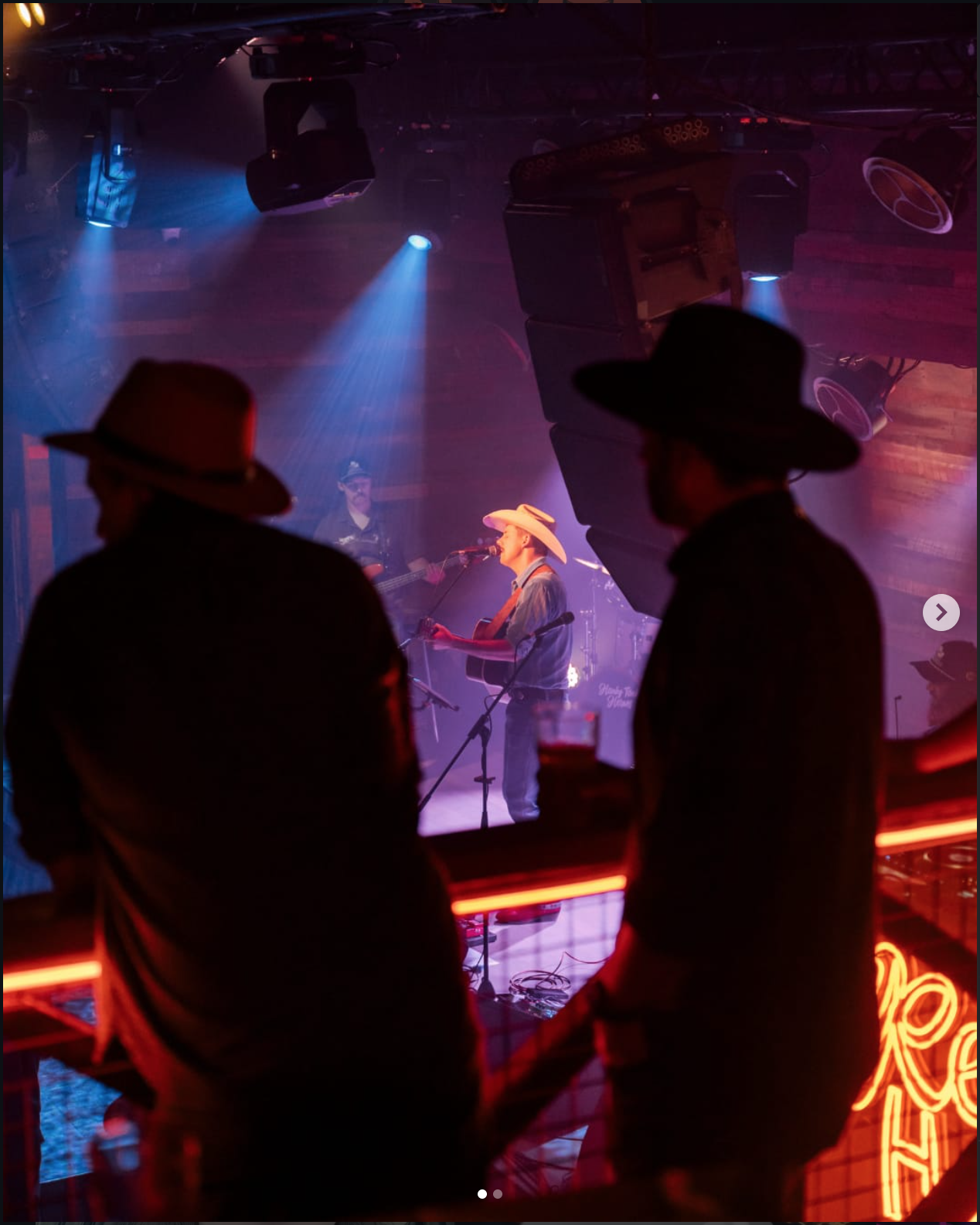 Live country music performance at a nightclub featuring a singer with a cowboy hat playing guitar, with band members in the background, viewed from a balcony with silhouettes of audience members.