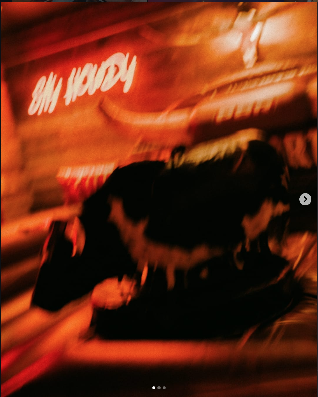 A black and white dog with a curly tail walking past a brightly lit red neon 'Krispy' sign, possibly in a bakery or doughnut shop, with a reflective surface creating a distorted view.