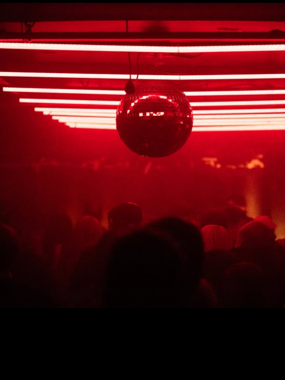 A dark club scene with red lighting and a large disco ball hanging from the ceiling, reflecting light onto the crowd below.