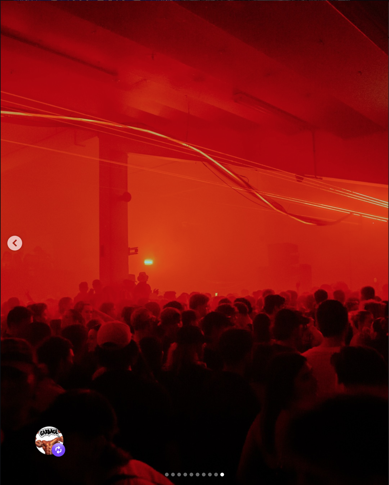 A large crowd of people in a dimly lit indoor space illuminated by red lights and laser-like rays, with a few people standing near a pillar and some standing in the background.