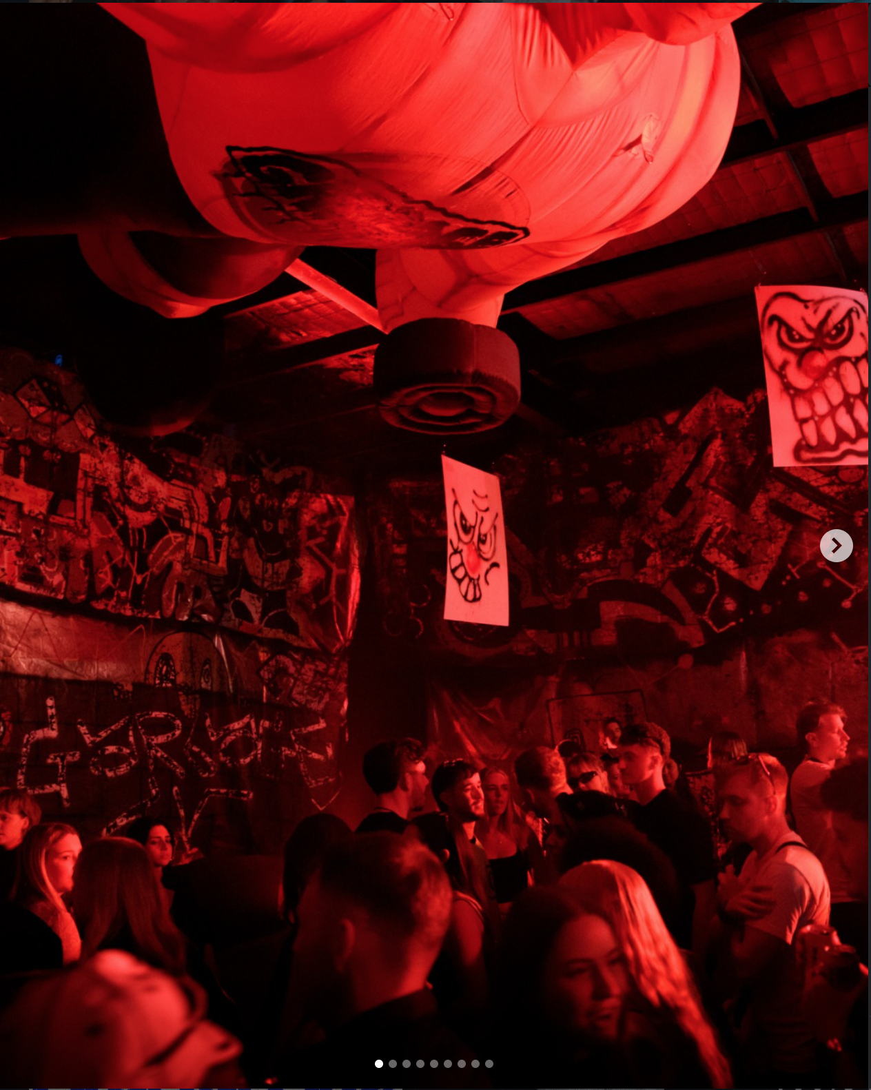 People gathered at a dark-themed party or event with red lighting, graffiti-style artwork, and decorative paper lanterns hanging from the ceiling.