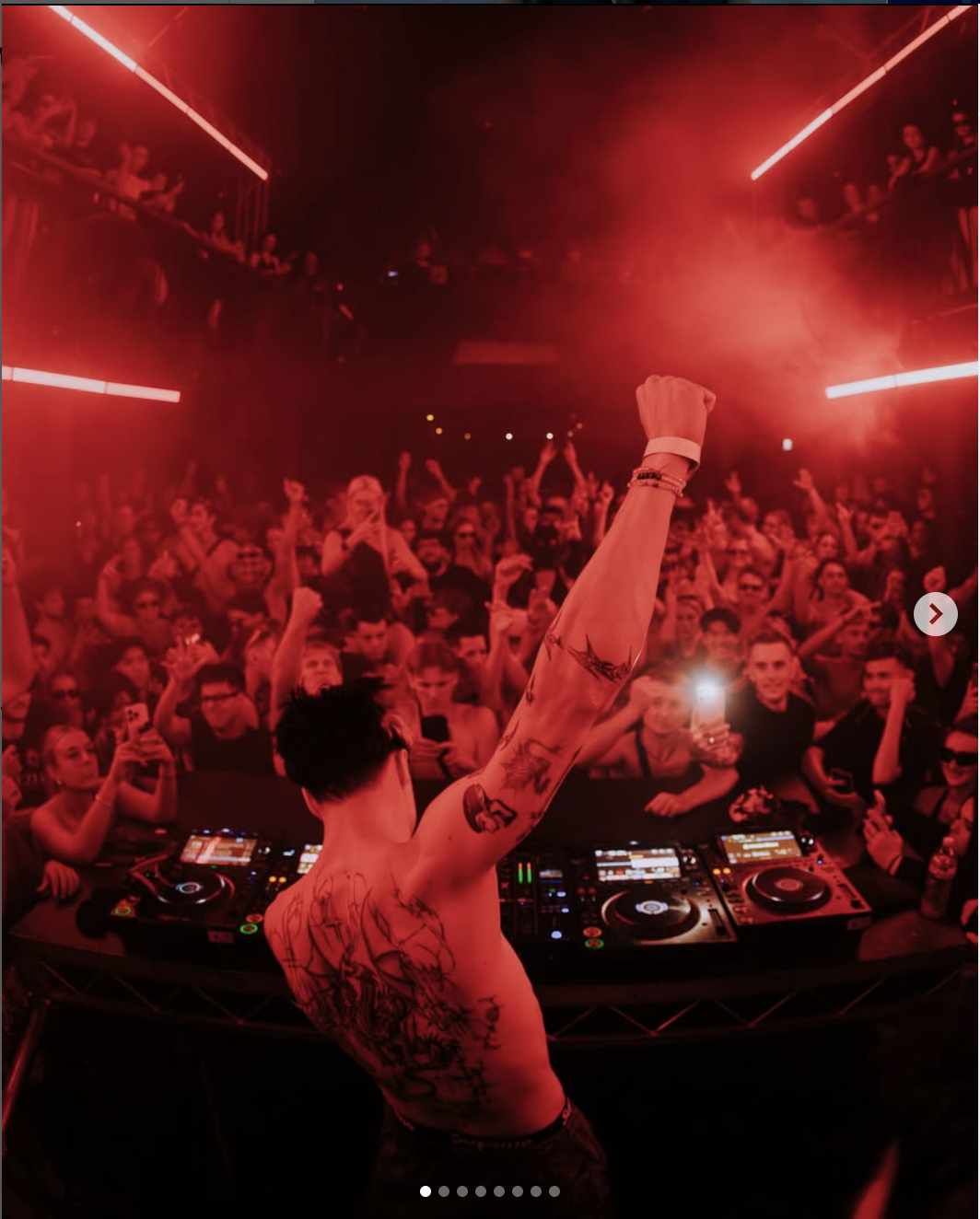 A DJ with tattoos on his back and arm raising his fist in front of a crowd at a nightclub, illuminated by red lights and laser beams, with people cheering and taking photos.