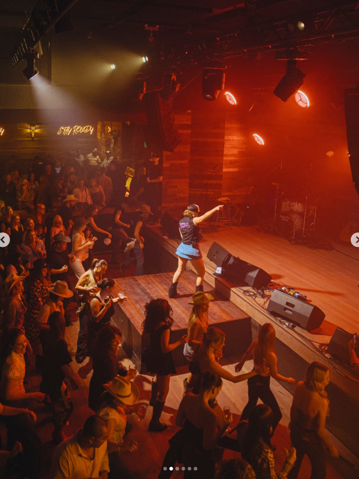 A lively country music concert at a bar or music venue with a stage, band, and audience dancing and enjoying the performance. The atmosphere is warm with red and yellow stage lighting.