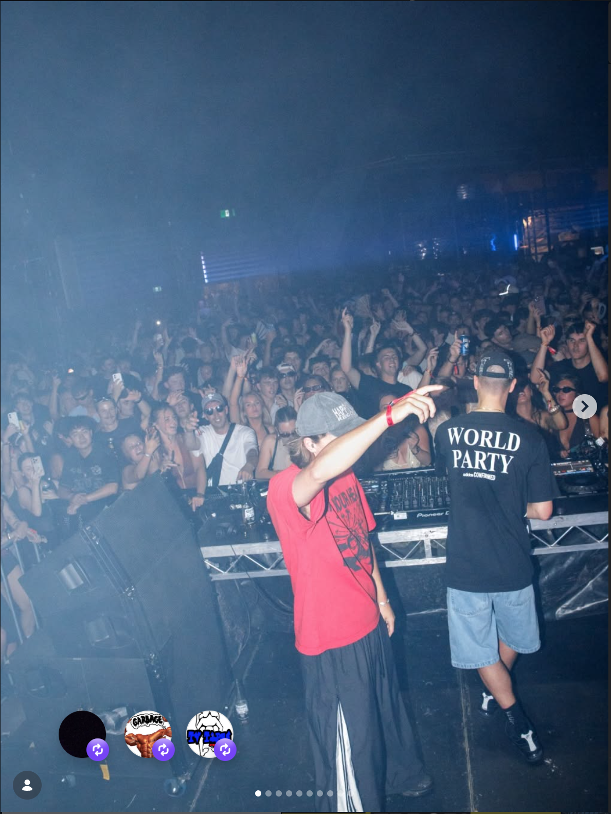 A crowded nightclub with a DJ on stage wearing a black T-shirt that says "WORLD PARTY." The audience is dancing and taking photos, with a woman in a red shirt and gray cap pointing towards the crowd.