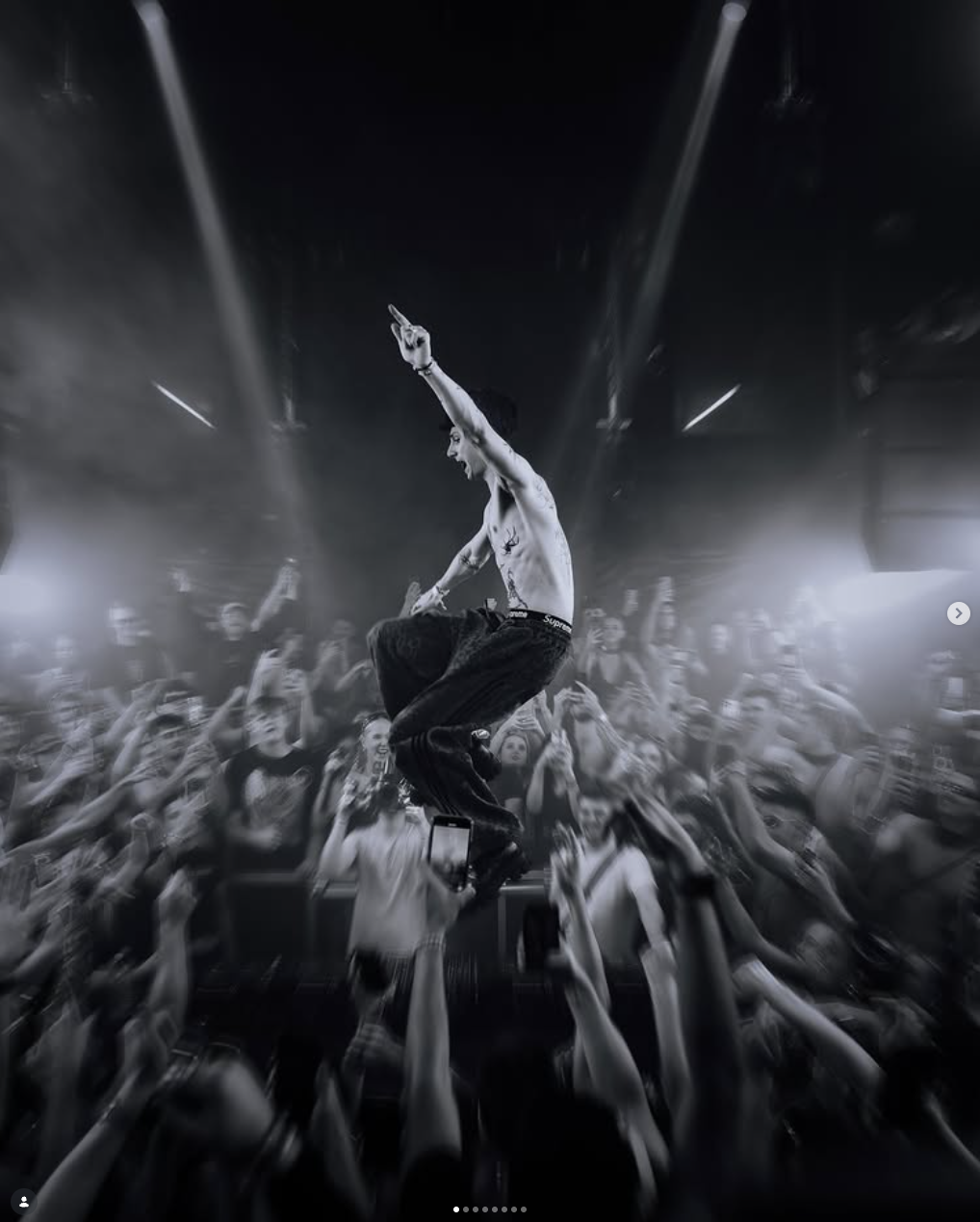 A shirtless male performer with tattoos, jumping amidst an excited crowd in a concert or nightclub, with dramatic lighting and a lively atmosphere.