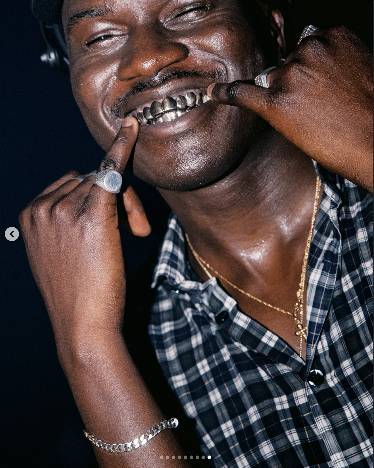 A close-up of a man with dark skin smiling and pointing to his gold teeth with his index fingers, wearing jewelry and a plaid shirt.