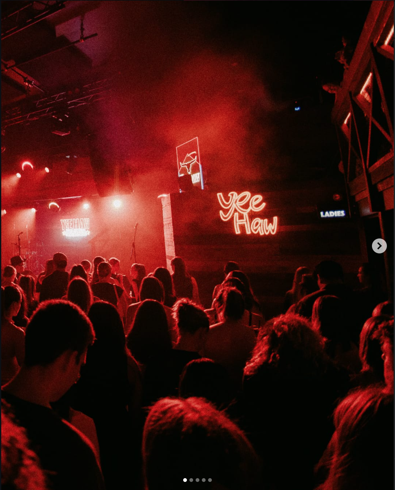 Crowd of people in a dimly lit concert venue with red lighting, stage, and neon signs that say 'Yee Haw' and 'Ladies'.
