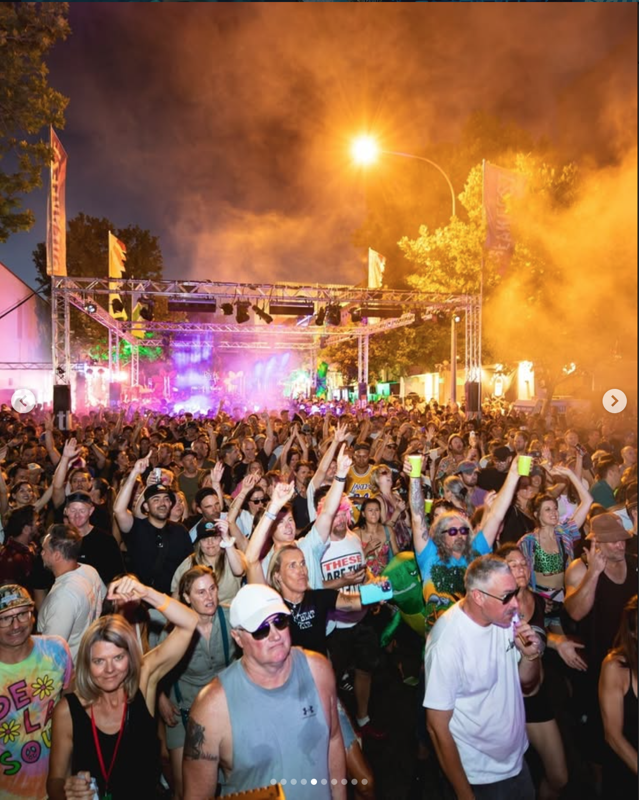 Crowd at a concert or festival outdoors at night with stage lighting and smoke effects, many people raising their hands, some holding drinks, and visible stage equipment under a bright streetlight.