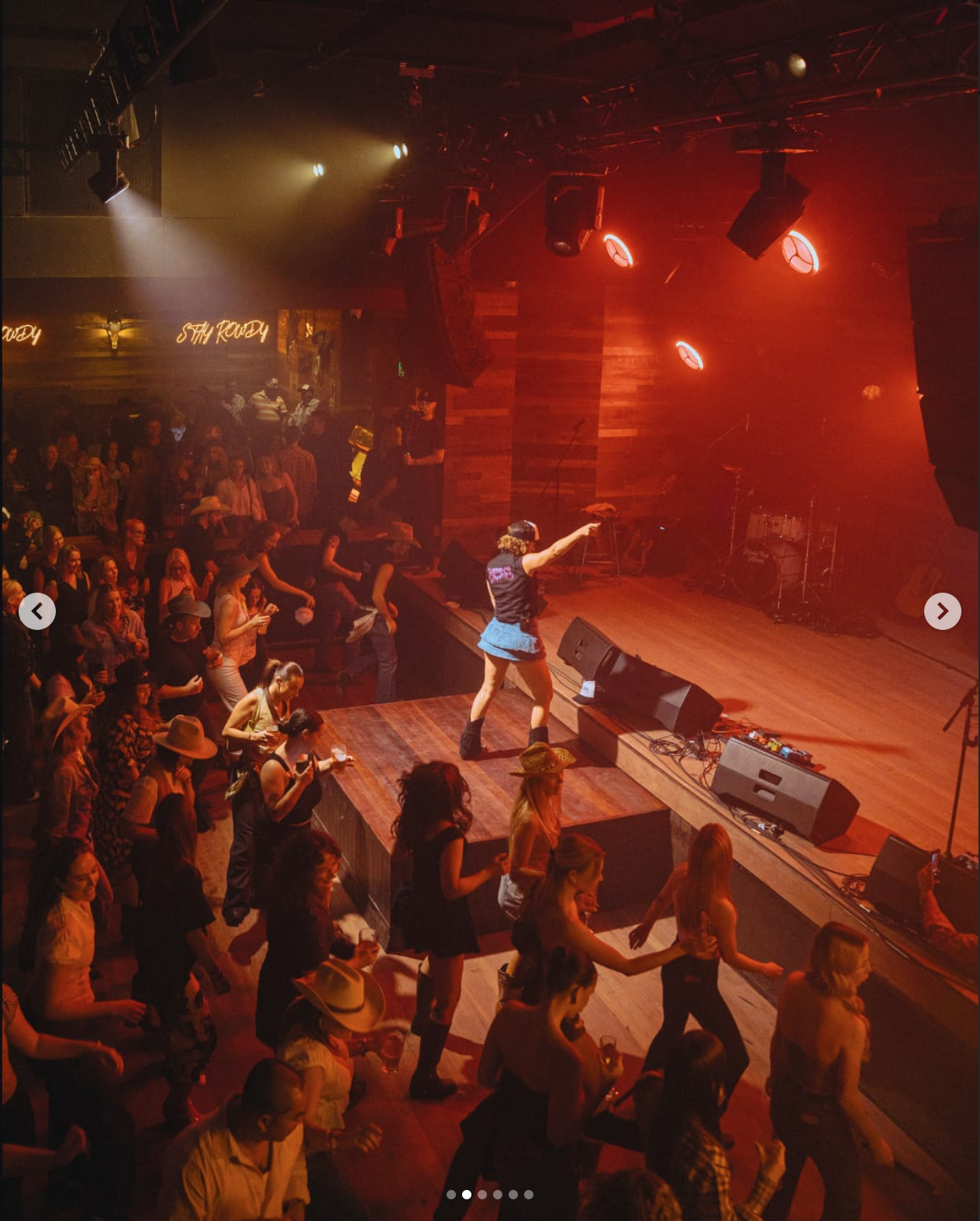 A lively concert or dance event with a diverse crowd of people dancing on the floor and some standing near the stage, where a person is performing or leading the dance. The stage has musical instruments and is lit with colorful lights, with warm, ambient lighting creating a festive atmosphere.