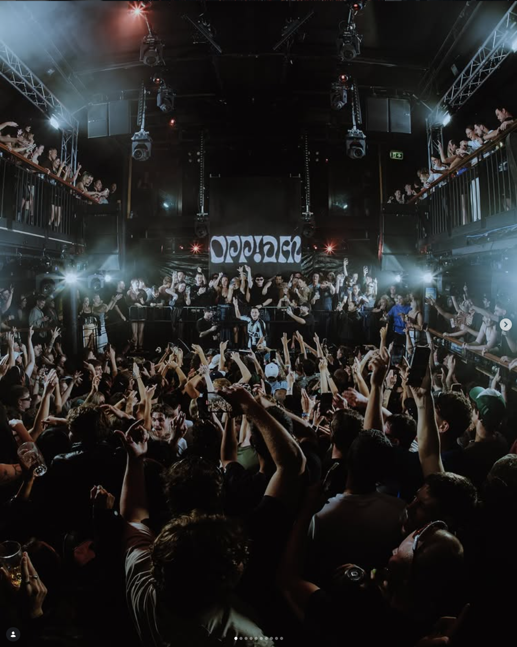 Crowded concert venue with a stage, crowd dancing and raising hands, and band performing on stage with a banner that says 'OPPIA'. Audience on the balcony and main floor, with bright concert lighting.