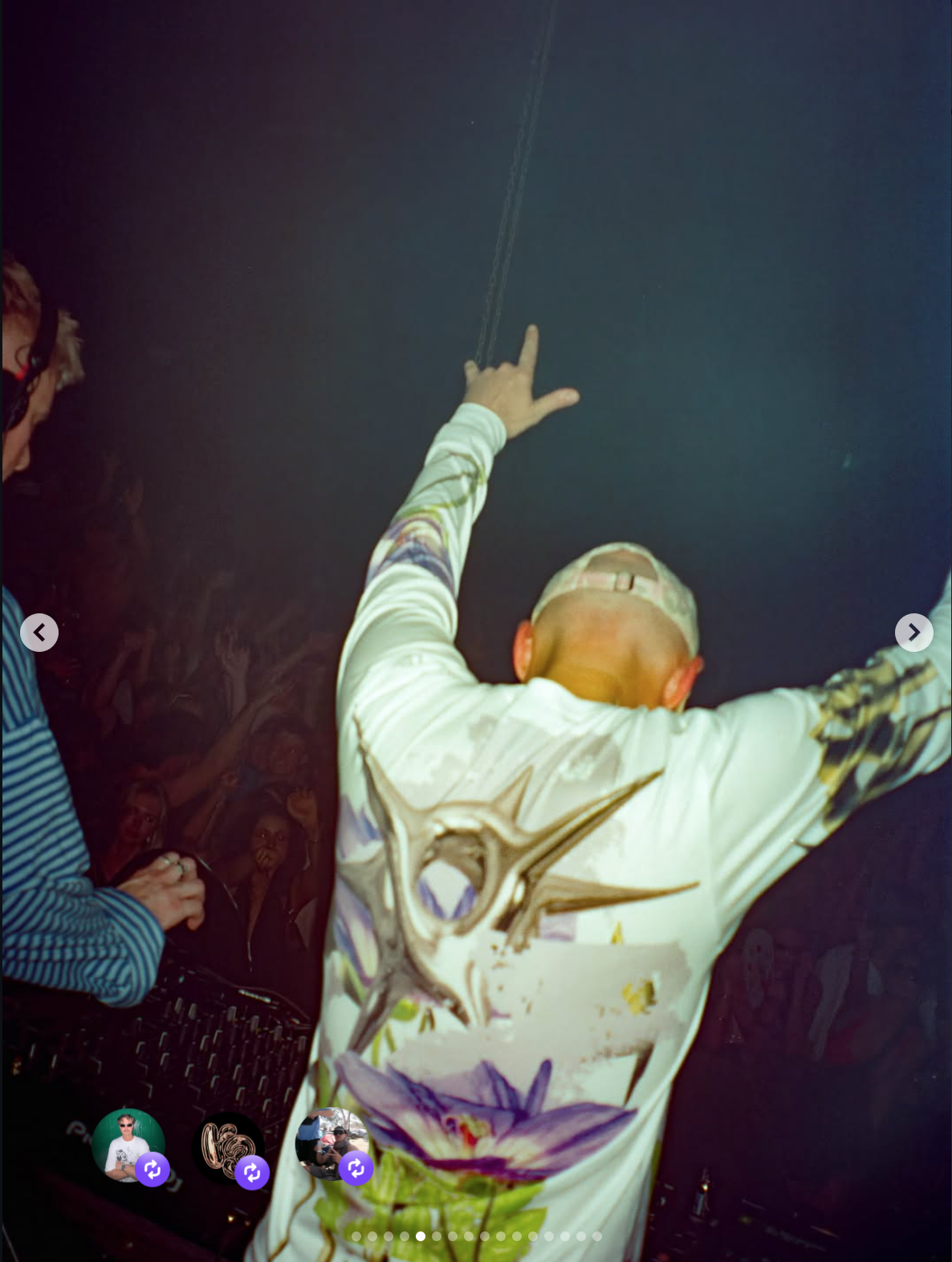 Person wearing glasses with a shaved head and tattoos, wearing a colorful jacket, raising hands in the air at a party or concert, with a DJ in the background and a crowd visible in the shadows.