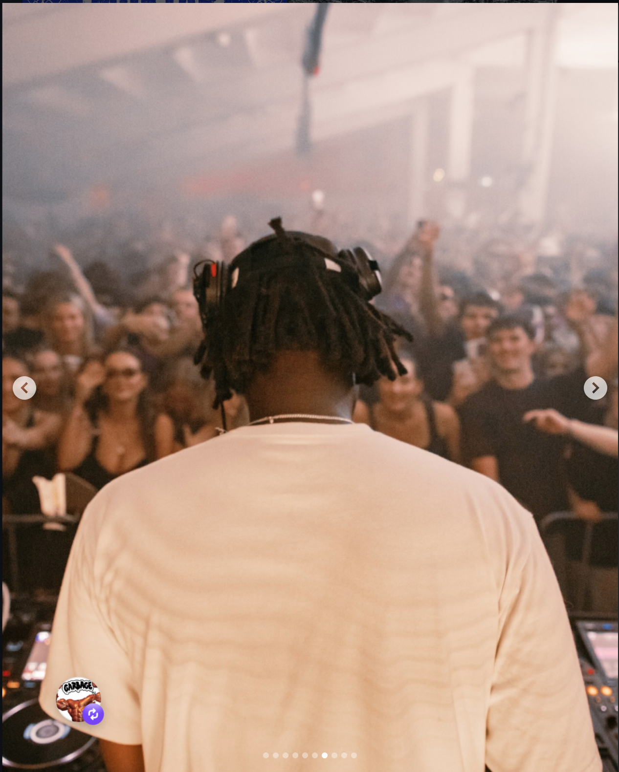A DJ with dreadlocks and headphones is seen from behind performing at a crowded nightclub or concert with people dancing and raising their hands in the background.