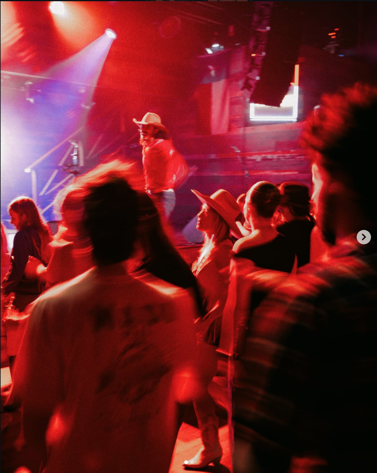 People dancing in a dimly lit nightclub with red and purple stage lighting, a performer wearing a cowboy hat on stage.