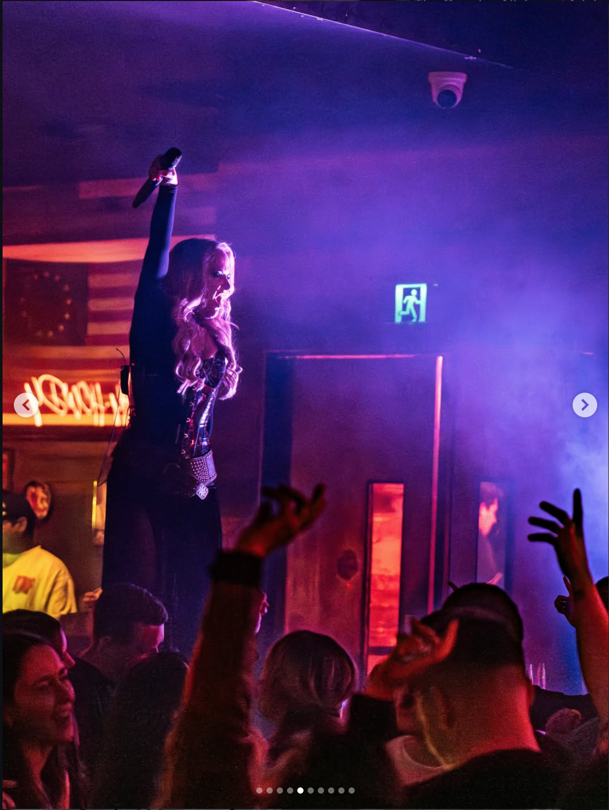 Woman performing on stage with microphone in hand, surrounded by a crowd at a concert or nightclub, illuminated by purple and red lights, with a neon sign and American flag in the background.