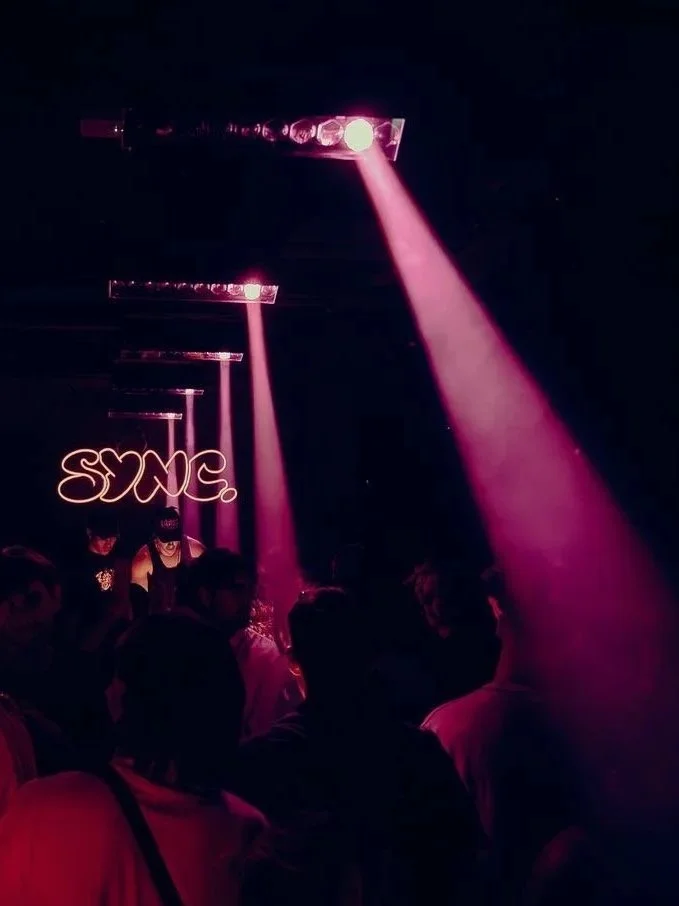 People dancing in a dark club with pink spotlight, neon sign reads I LOVE DANCE.