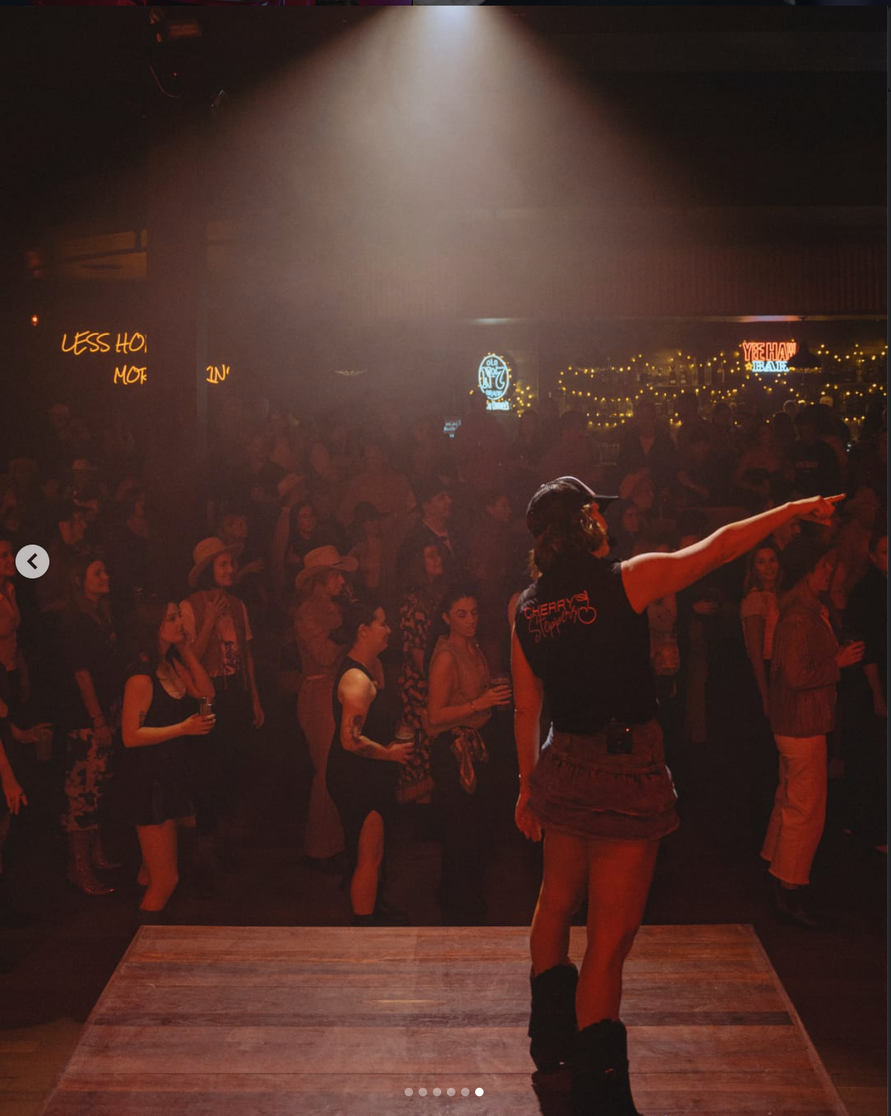 A woman on stage pointing with her right arm at a crowded dance hall or concert venue. The audience is engaged, some holding drinks, and there are neon signs and string lights in the background.