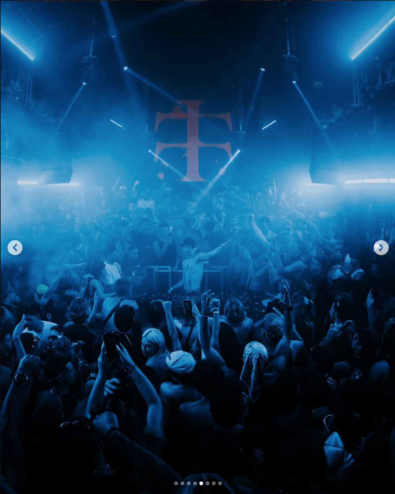 A crowded nightclub with a DJ performing on stage under blue lighting and fog effects, with people dancing and taking photos.