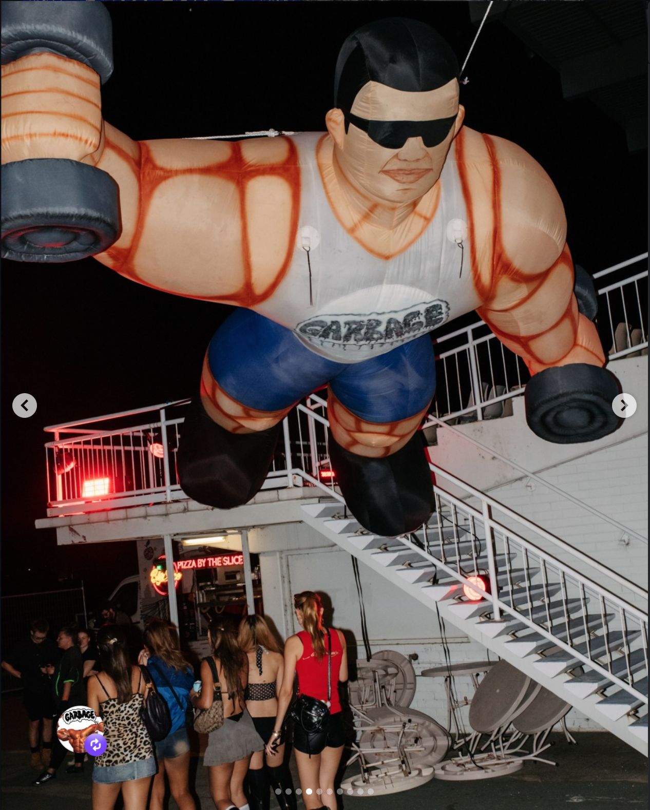 A large inflatable figure of a muscular man wearing sunglasses, a white tank top with a logo, blue shorts, and black shoes, hanging above a group of people outside a building at night.