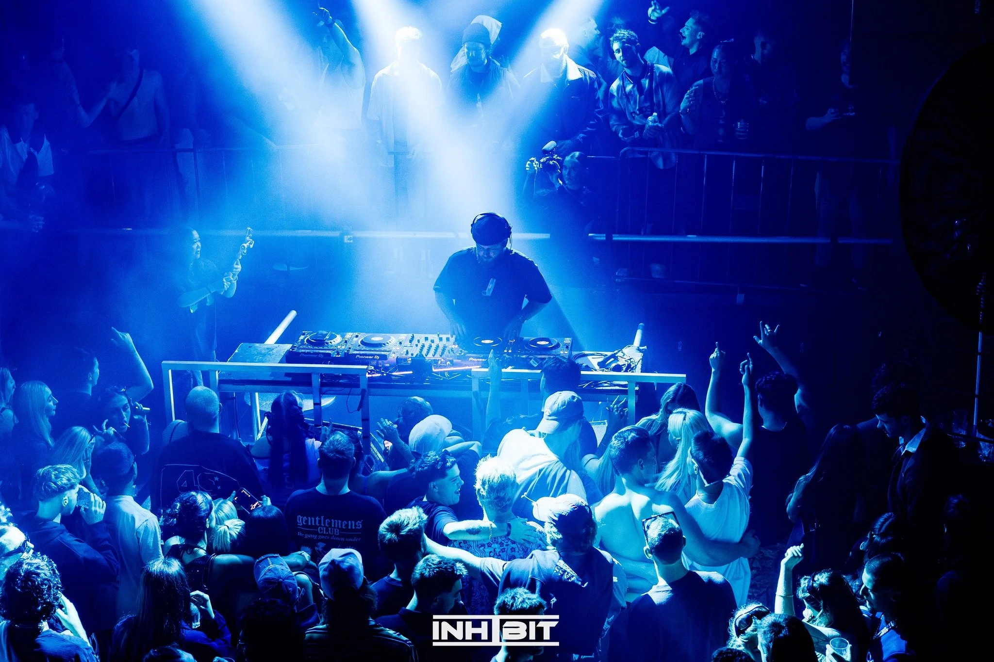 A DJ performs at a nightclub surrounded by a large crowd of dancing people, with blue lighting and an 'INHIBIT' logo at the bottom.