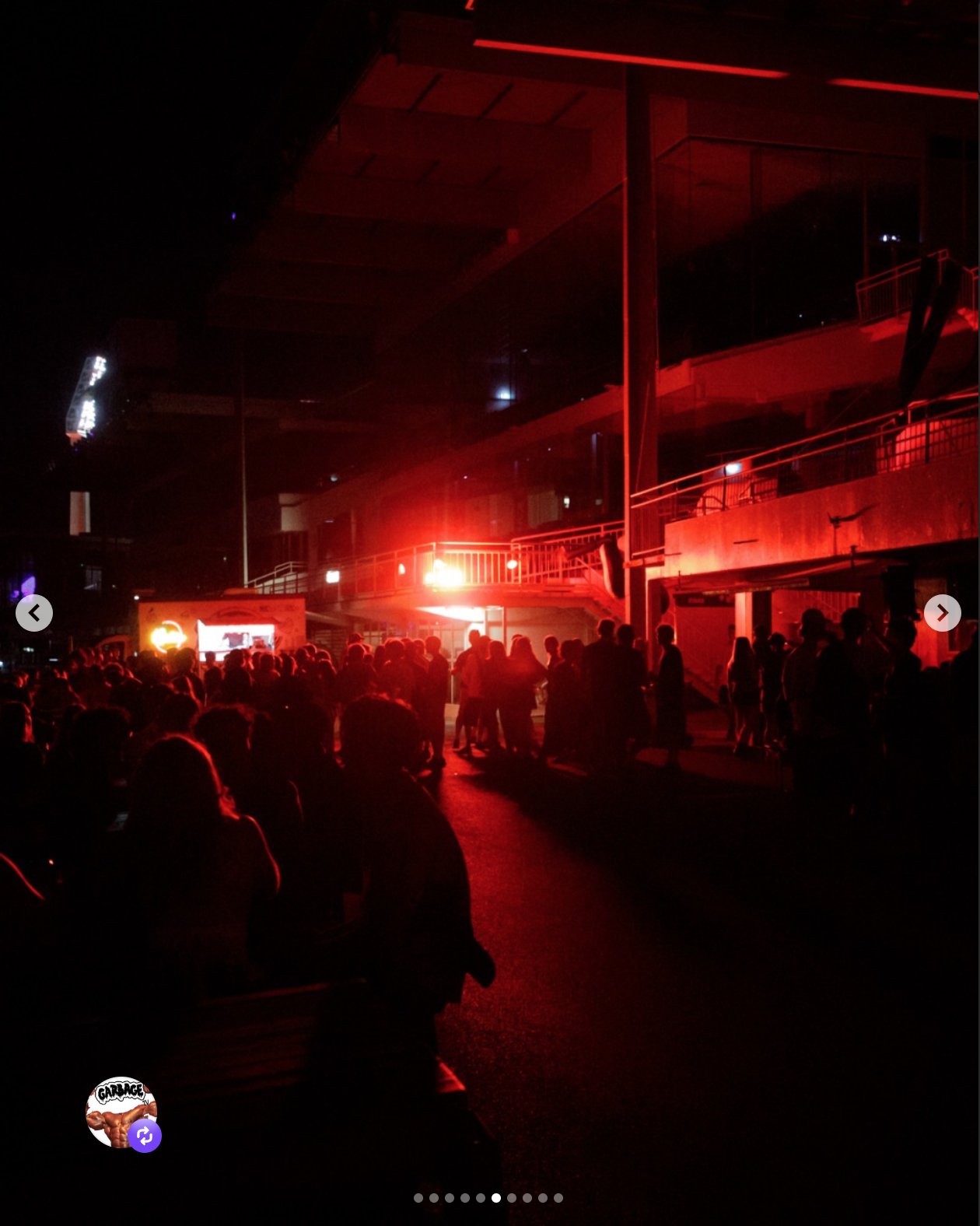 Crowd of people gathered outside a building at night, illuminated by red lights and possibly a neon sign, with a large screen in the background.