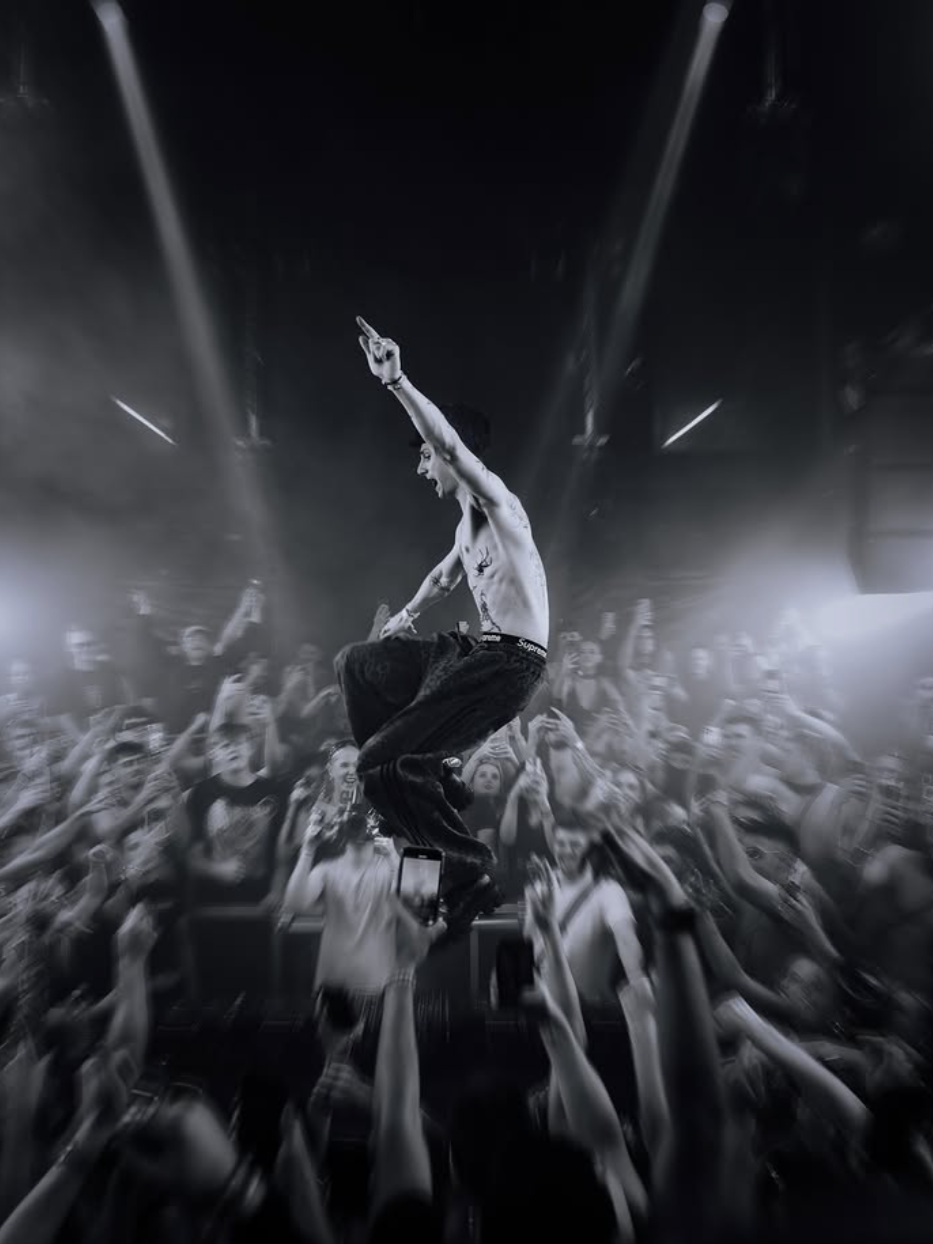 A shirtless performer in black pants jumps on stage with arms raised, surrounded by a crowd at a concert or nightclub, black and white photo.