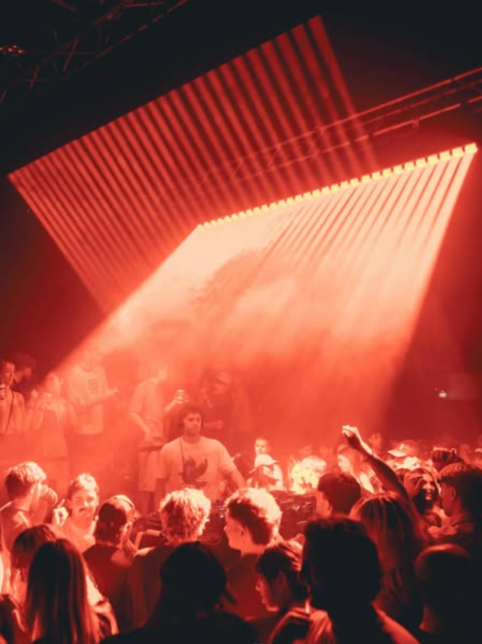 Crowd of people dancing and socializing at a club or concert with red stage lighting and large overhead LED screens.