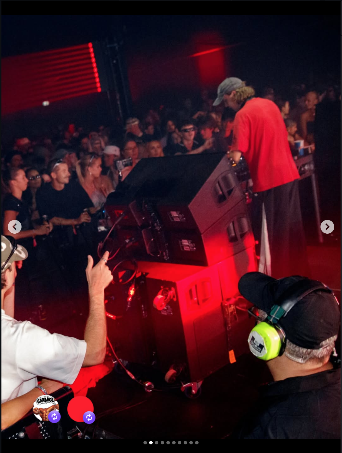 A DJ in a red shirt and gray hat performs at a crowded indoor music event with red lighting, with people in the audience enjoying the show and various equipment around him.