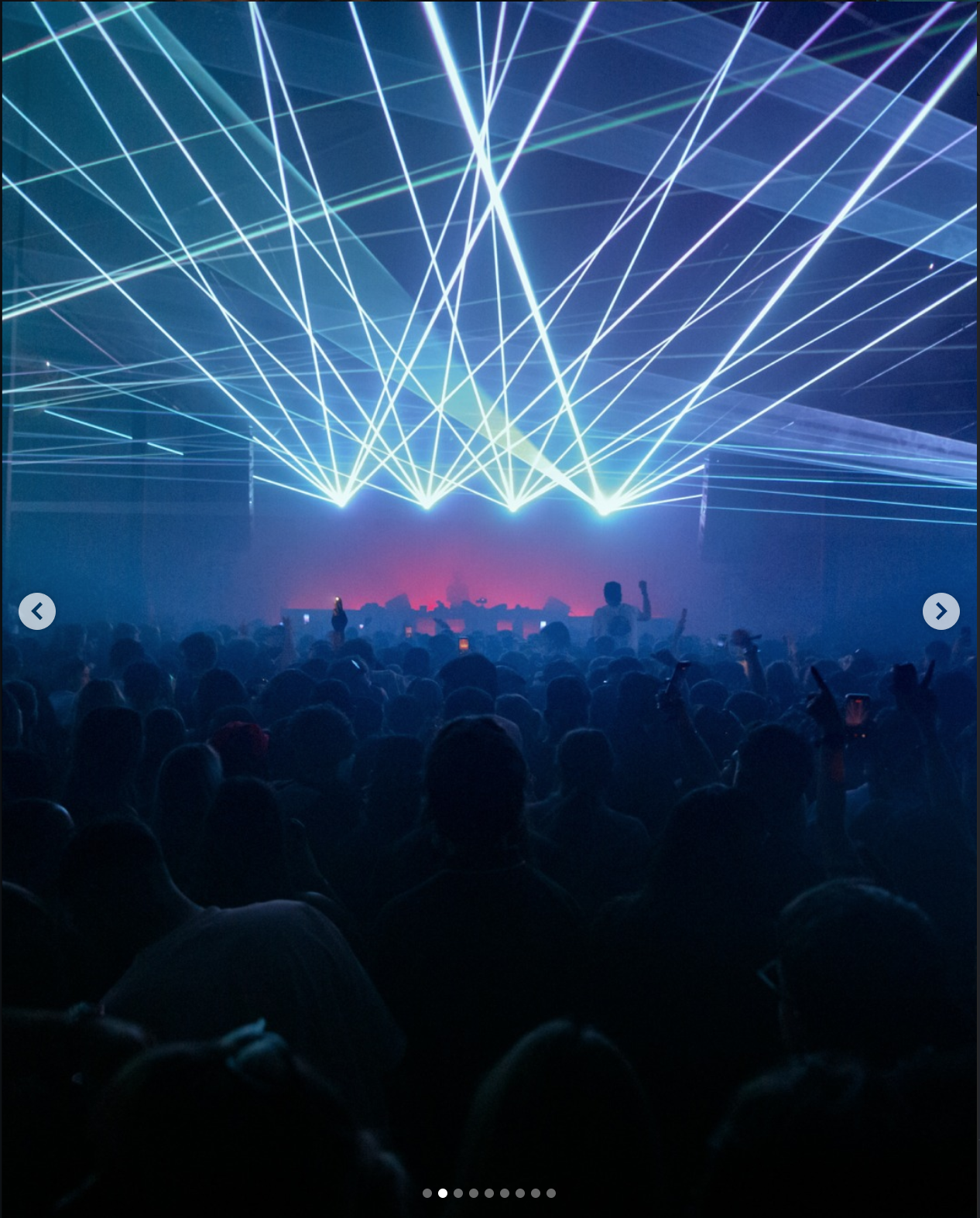 A concert scene with a large crowd facing a stage, illuminated by multiple bright laser lights projecting in various directions across the dark venue.