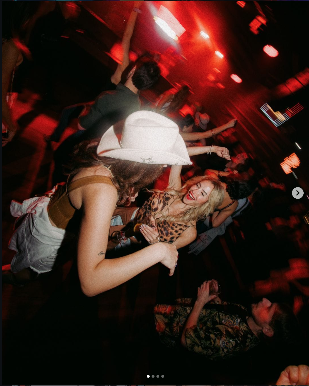 People dancing and socializing in a dimly lit nightclub with red lighting, including a woman with blonde hair wearing a leopard print top and a woman with a white cowboy hat and light-colored outfit.