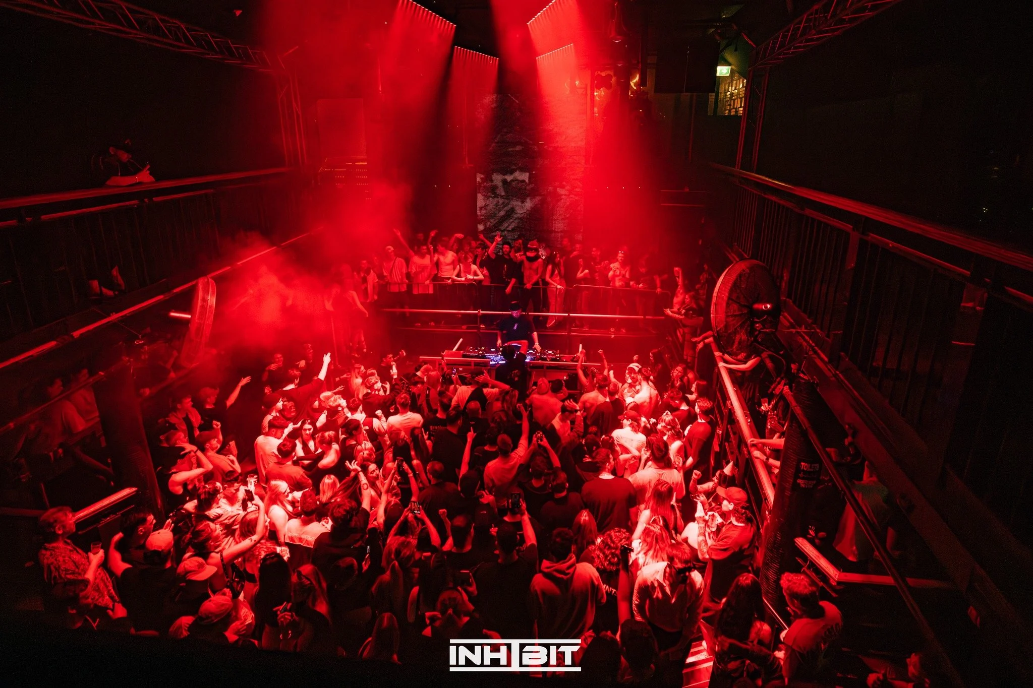 Crowd of people dancing and enjoying a DJ performance in a dark nightclub with red lighting, fog effects, and a DJ on stage. The nightclub has a multi-level design with railings and a large banner displaying the word 'INHIBIT' at the bottom center.