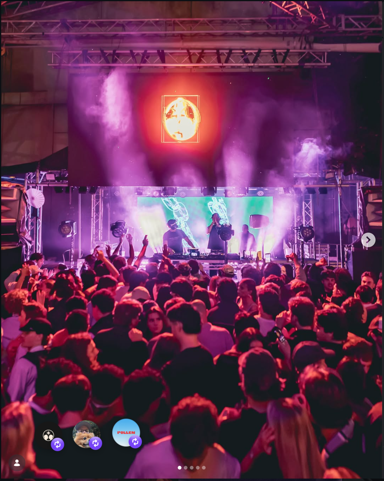 A large crowd at a concert or music festival with a DJ performing on stage. The stage has colorful lighting, smoke effects, and a vibrant digital screen with graphics. The audience is engaged, with many raising their hands and dancing.