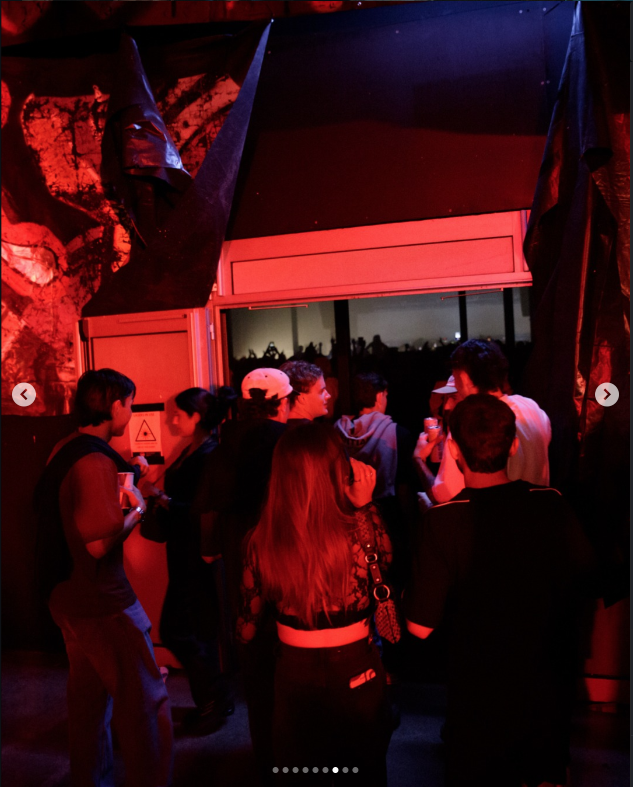 Group of people in a dimly lit room with red and purple lighting, some wearing casual clothes and hats, standing and socializing near a large window.