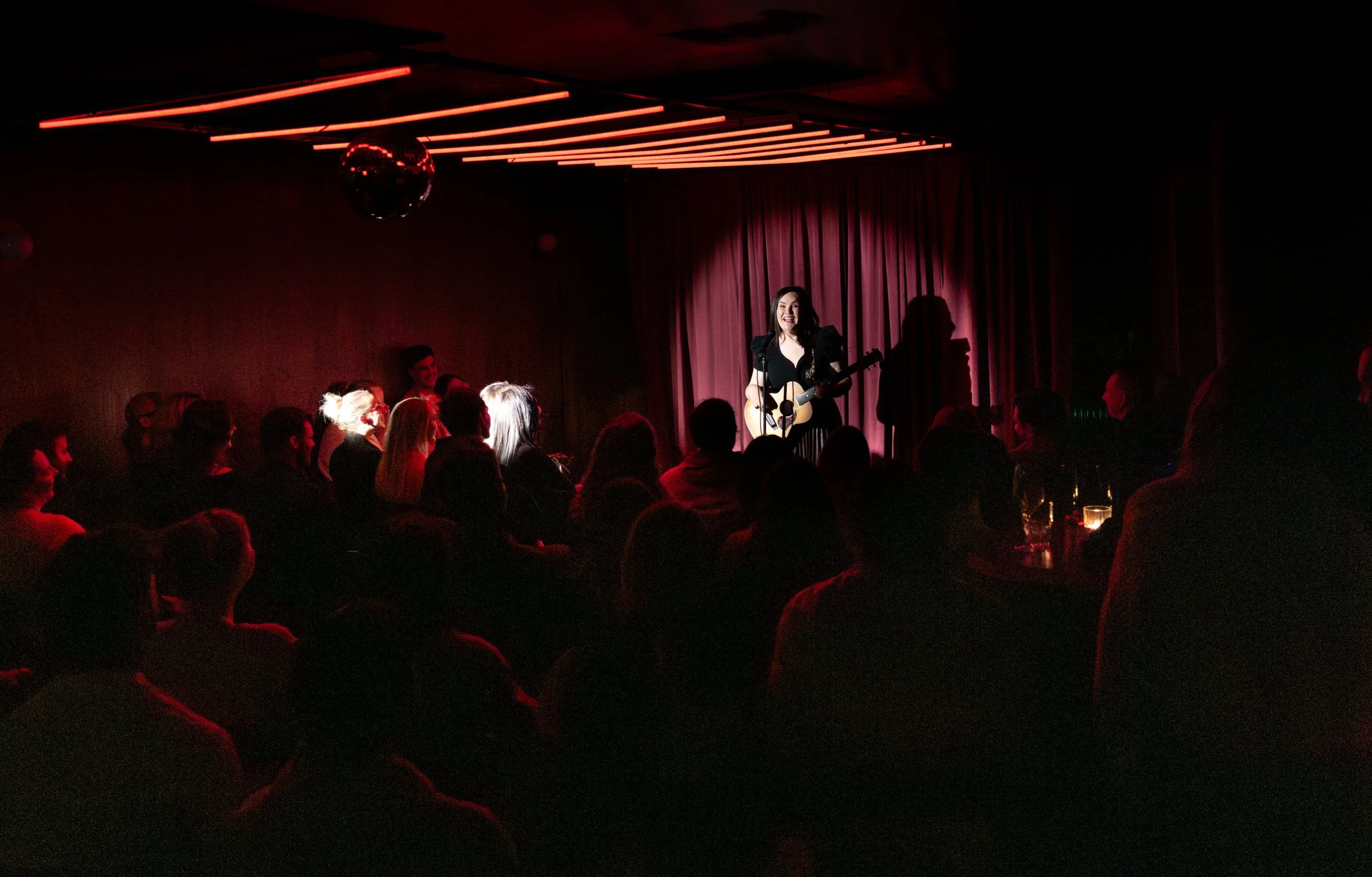 A woman singing and playing guitar on a small stage, performing for an audience in a dimly lit venue with red lighting and decorated ceiling panels.