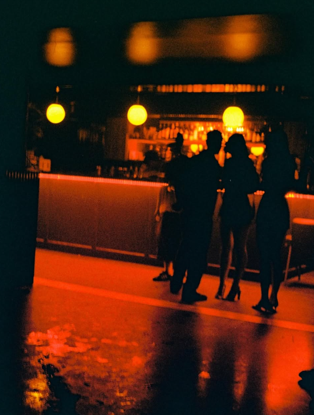 Silhouettes of people at a dimly lit bar with warm orange lighting, bar shelves in the background, and reflections on the floor.