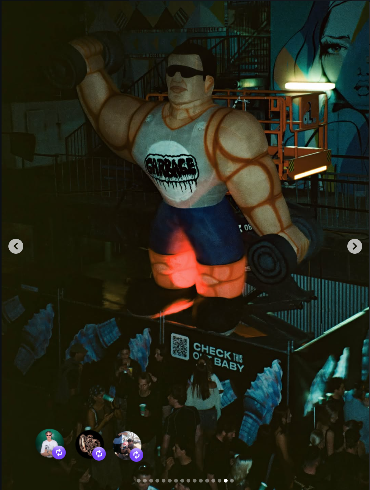 A large animated figure of a muscular man with dark hair, wearing sunglasses, a gray tank top with a design that says 'Garbage,' orange shorts, and black sneakers. The figure is holding a rolled-up mat or letter in one hand and a dumbbell in the other. The scene appears to be from an amusement park or arcade, with people below and colorful murals and lighting in the background.